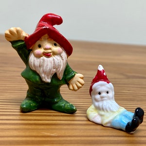 May include: Two miniature gnome figurines. The larger gnome stands wearing a dark green outfit and a red pointed hat with one arm raised. The smaller gnome is in a reclining position, wearing a red hat, a pale yellow shirt, and blue pants.