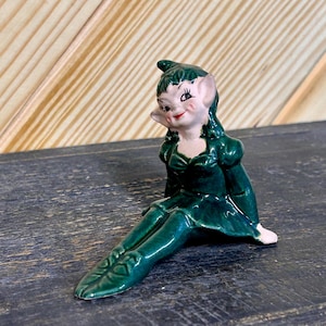 May include: A ceramic figurine of a seated elf with a green dress and pointed hat. The elf has a smiling face with rosy cheeks and red lips. The figurine is on a dark surface with a wooden background.