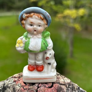 May include: A vintage porcelain figurine of a young child wearing a blue hat and green vest, holding a basket of yellow flowers and standing with a small dog. The figurine has red shoes and is on a white base.