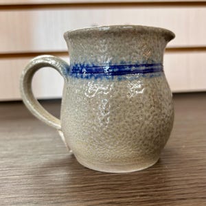 May include: A small, handmade stoneware pitcher with a textured, speckled surface. The pitcher is primarily beige with a blue band around the middle. It has a curved handle and a small spout. The pitcher is likely used for serving beverages.
