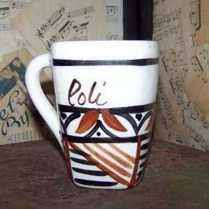 May include: White ceramic mug with a curved handle, featuring a black and brown geometric design and the name "Poli" in brown script. The mug has a black stripe near the rim.