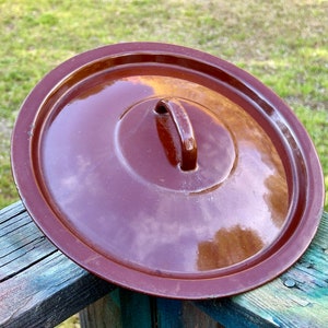 May include: A round, brown ceramic lid with a handle. The lid has a glossy finish and a slightly raised rim. The handle is a small, curved loop. The lid is likely for a pot or casserole dish.
