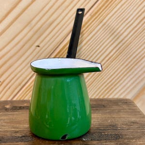 May include: A small, green enamelware pitcher with a black handle and spout. The spout is white-rimmed. The pitcher is set on a wooden surface. The pitcher is likely used for pouring liquids.