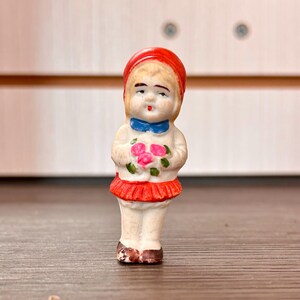 May include: A small, vintage porcelain figurine of a girl holding pink roses. The figurine wears a red bonnet, a blue bow, and a red skirt. The doll is painted with delicate features and stands on a wooden surface.