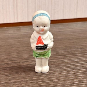 May include: A small porcelain figurine of a child with a light blue headband, wearing a white collared shirt and green shorts. The figure is holding a small, triangular slice of watermelon with a red top and a black rind in its hands.