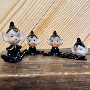 May include: Four vintage ceramic elf figurines with black outfits and hats, white faces, and rosy cheeks. The elves are in a seated position with their legs outstretched. The figurines are displayed on a wooden surface.