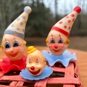 May include: Three vintage clown figurines with painted faces, red noses, and colorful hats. Two clowns wear cone-shaped hats with polka dots, and one wears a small yellow hat. They are set on a red woven base.