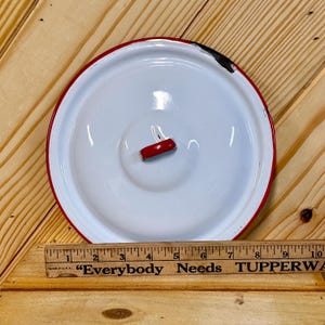 May include: A white enamel lid with a red rim and a red handle. The lid is circular and has a slightly raised center. The image also shows a wooden ruler with the text "Everybody Needs".