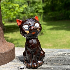 May include: A glossy, brown ceramic cat figurine with orange-tipped ears and white eyes. The vintage cat is in a seated position, with a slightly open mouth and a gold accent on its face. The figurine is on a weathered wooden surface.