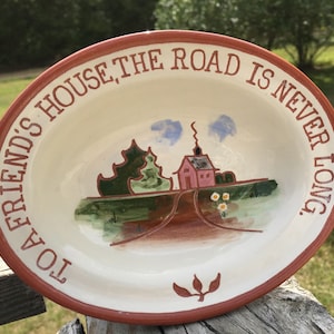 May include: Oval ceramic serving dish with a cream-colored interior and a reddish-brown rim. The dish features a painted scene of a house and road, with the text "TO A FRIEND'S HOUSE, THE ROAD IS NEVER LONG."