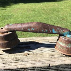 May include: An antique, rusty metal object with a curved handle and two cylindrical bases. The metal has a weathered appearance with patches of turquoise paint, suggesting age and outdoor use. The object sits on a weathered wooden surface.