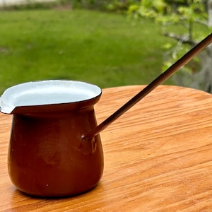 May include: A small, brown enamel coffee pot with a long, thin handle. The interior and rim are white. The pot is sitting on a wooden surface with a green background.