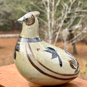 May include: A ceramic bird figurine with a cream-colored body and brown and blue painted details. The bird has a long neck, a small beak, and a decorative pattern of birds and floral designs. The figurine is sitting on a wooden surface.