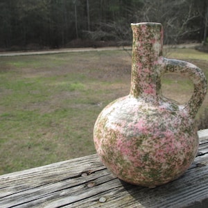 May include: A speckled ceramic pitcher with a round base, a tall neck, and a handle. The pitcher is primarily pink and green, with a white interior. The pitcher is sitting on a wooden surface.