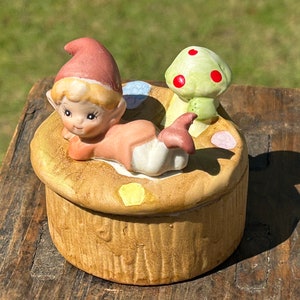 May include: A ceramic trinket box featuring a whimsical design. The box is shaped like a mushroom with a small elf figurine and a green mushroom with red spots on top. The elf wears a pink hat and is lying down.