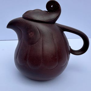 May include: A dark brown ceramic teapot with a unique design. The teapot features a curved spout, a handle, and a lid with a decorative swirl. The body of the teapot has a textured, ribbed pattern.