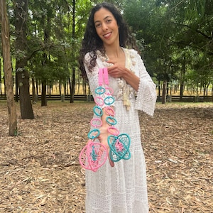May include: Two decorative hanging ornaments featuring interconnected pink and teal plastic rings. Each ornament ends in a spherical, cage-like structure woven with matching pink and teal thread, suspended from a bright pink fabric strap.