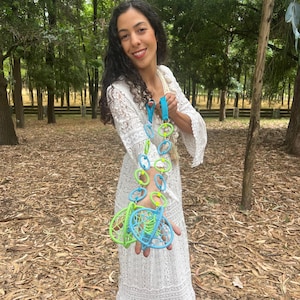 May include: A long decorative hanging ornament featuring a series of interconnected light blue and lime green circular rings, ending in two spherical woven structures made of matching colored cord, suspended from a bright blue fabric strap.
