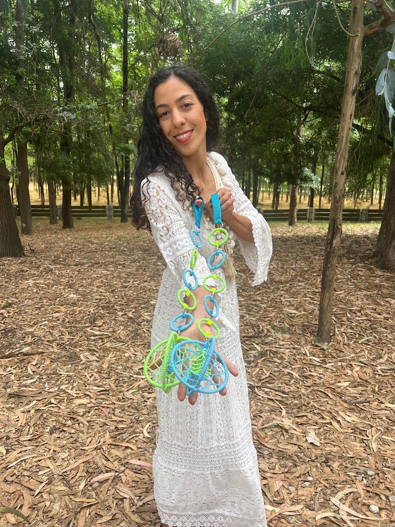 May include: A long decorative hanging ornament featuring a series of interconnected light blue and lime green circular rings, ending in two spherical woven structures made of matching colored cord, suspended from a bright blue fabric strap.