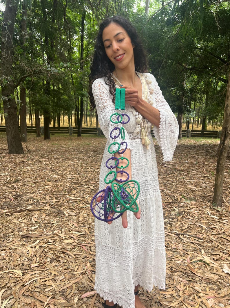May include: A decorative hanging ornament featuring a vertical chain of alternating purple and green circular rings connected by silver metal jump rings, ending in two larger, spherical, webbed structures in matching purple and green yarn.