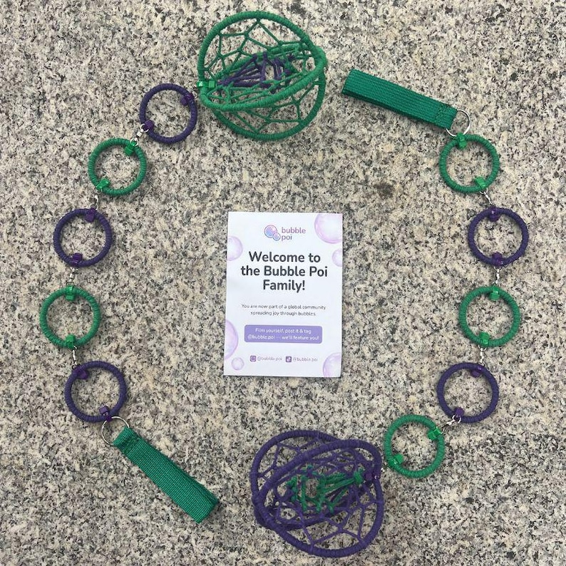 May include: A bubble poi set featuring two spherical, woven net heads in green and purple, connected by a chain of alternating green and purple rings. Two green fabric finger loops are attached to the ends. A white card reads Welcome to the Bubble Poi Family.