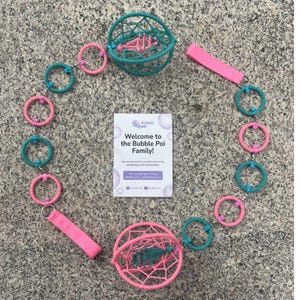 May include: A bubble poi set featuring two spherical cages, one teal and one pink, connected by a chain of alternating teal and pink rings. Two pink fabric finger loops are attached to the ends. A white card reads Welcome to the Bubble Poi Family.