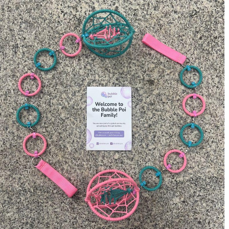 May include: A bubble poi set featuring two spherical cages, one teal and one pink, connected by a chain of alternating teal and pink rings. Two pink fabric finger loops are attached to the ends. A white card reads Welcome to the Bubble Poi Family.