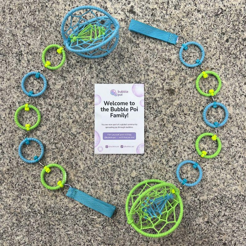 May include: A bubble poi set featuring two spherical, webbed heads in light blue and lime green, connected by a chain of alternating light blue and lime green wrapped rings. Two light blue fabric handles are attached. Includes a white card with the text: Welcome to the Bubble Poi Family! You are now part of a global community spreading joy through bubbles.