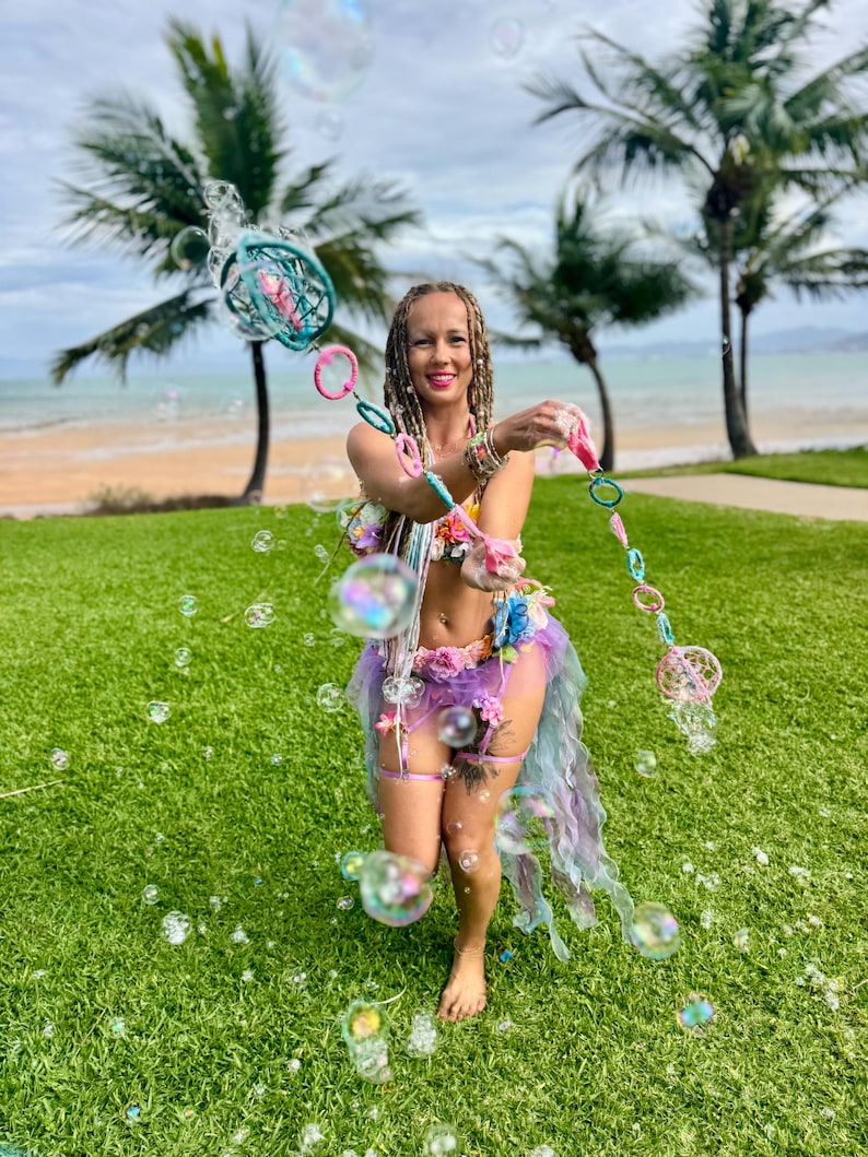 May include: A bubble wand featuring a series of interconnected circular frames in shades of teal, pink, and blue, designed for creating large bubbles. The wand is held horizontally, showcasing its colorful, lightweight structure and circular loops.