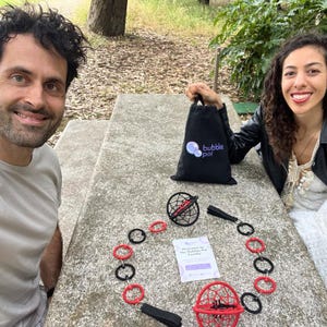 May include: A black drawstring bag with the white and blue text bubble poi, a poi set featuring two spherical cages in red and black, connected by a chain of alternating red and black rings, and a white informational card with the text Welcome to the Bubble Poi Family.