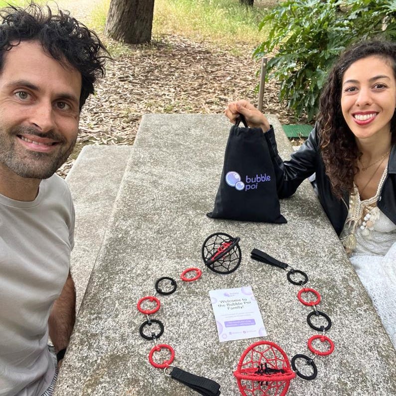 May include: A black drawstring bag with the white and blue text bubble poi, a poi set featuring two spherical cages in red and black, connected by a chain of alternating red and black rings, and a white informational card with the text Welcome to the Bubble Poi Family.