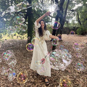 May include: A long, cream-colored dress with intricate lace detailing and three-quarter length sleeves, worn while holding a bubble wand that creates a large, iridescent cloud of floating soap bubbles in an outdoor setting.
