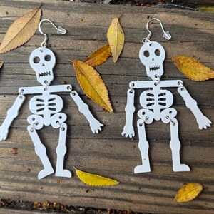 May include: A pair of white skeleton earrings with silver accents. The skeletons are articulated at the joints, giving them a movable appearance.