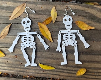 May include: A pair of white skeleton earrings with silver accents. The skeletons are articulated at the joints, giving them a movable appearance.