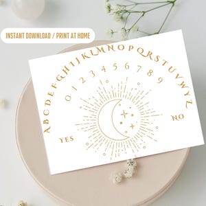 May include: A rectangular white card featuring a gold-colored arc of letters from A to Z, numbers 0 through 9, and the words YES and NO. In the center, a gold-colored crescent moon is surrounded by radiating lines and small star shapes.