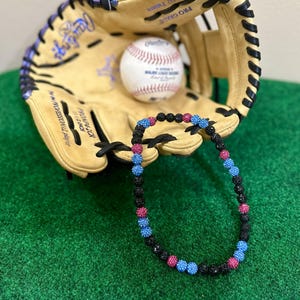 May include: A baseball glove and baseball rest on a green surface. A black beaded necklace with pink and blue accents is in front of the glove. The baseball is white with red stitching and the glove is tan.