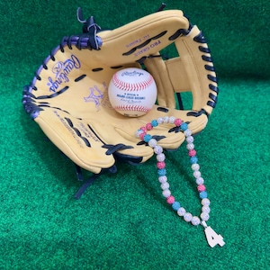 May include: A baseball glove with a baseball inside and a multi-colored beaded necklace with a number 4 charm hanging from it. The glove is tan with black stitching and a purple trim. The baseball is white with red stitching and the words "Rawlings Official Major League Baseball" printed on it. The necklace is made of white, pink, and turquoise beads.