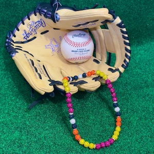May include: A baseball glove and baseball rest on a green surface. The baseball is white with red stitching and the Rawlings logo. A colorful beaded necklace with yellow, orange, pink, white, and navy beads is draped across the glove.