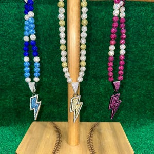 May include: Three silver chains with a lightning bolt pendant. The chains are made of small, round beads that are blue, gold, and pink. The chains are hanging from a wooden stand with the text "G-MONEY" on it.