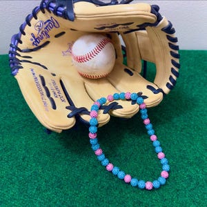 May include: A baseball glove with a pink and blue beaded necklace resting on it. The glove is brown with a purple and blue trim. The baseball is white with red stitching.