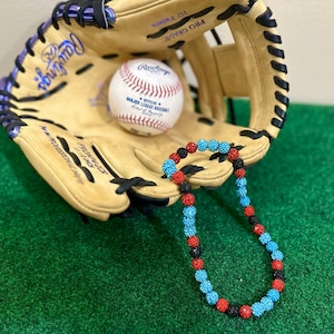May include: A baseball glove with a baseball and a beaded necklace. The baseball is white with red stitching and the Rawlings logo. The necklace has blue, red, and black beads. The glove is tan with purple accents.