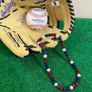 May include: A red, white, and blue beaded necklace with a baseball theme. The necklace is lying on a green artificial grass surface next to a baseball glove and a baseball.