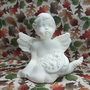 Könnte beinhalten: Eine weiße Keramik-Cherub-Figur mit detaillierten Flügeln, die einen Korb mit Trauben hält. Der Cherub sitzt mit einem ausgestreckten Bein. Der Hintergrund zeigt ein Muster aus Herbstblättern.