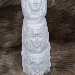 May include: A white, three-tiered sculpture featuring carved faces and headdresses. The top tier has horns and a circular emblem. The sculpture is set against a brown, textured background, suggesting a Native American theme.