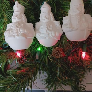 May include: Three white ceramic nutcracker ornaments hanging on a Christmas tree. Each ornament has a unique hat and uniform design. The ornaments are surrounded by green pine needles, red and green lights, and a pine cone.