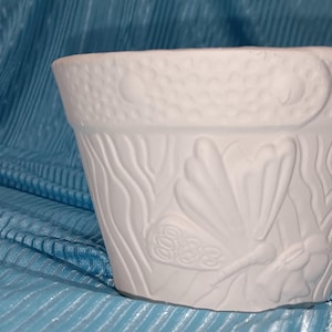 May include: A white ceramic flower pot with embossed seashell and seaweed designs. The pot has a textured rim and a slightly flared shape. The background is a blue fabric.