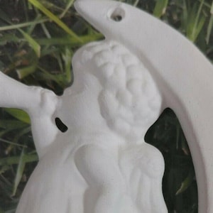 May include: A white ceramic angel figurine playing a horn, with a crescent moon-shaped accent. The angel has detailed facial features and curly hair. The background is blurred green foliage.
