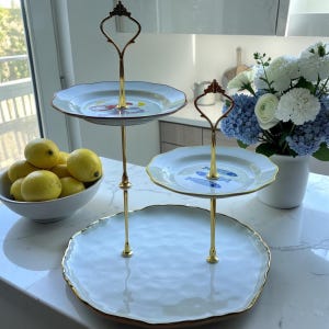 May include: A tiered serving stand featuring three white scalloped plates with gold-tone metal hardware. The top plate displays a colorful fish graphic, the middle plate shows blue fish illustrations, and the large base plate is plain white.