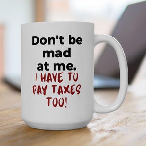 Può includere: Tazza in ceramica bianca con la scritta "Don't be mad at me. I HAVE TO PAY TAXES TOO!" in nero e rosso. La tazza ha un manico grande ed è appoggiata su una superficie di legno.