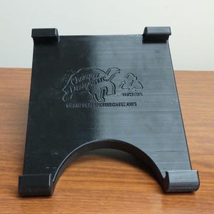 May include: Black plastic stand with a curved base and two side supports. The stand has a logo that says "Charger Designs Inc." with a turtle graphic and the text "Others" and "www.chargerdesignsinc.com".